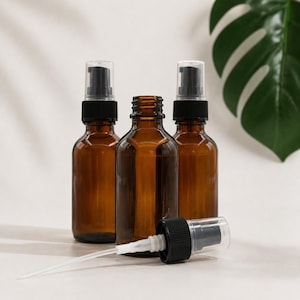 May include: Three amber glass bottles with black spray pump tops. Two bottles stand upright with pumps attached, while one central bottle is open without a cap. A single black spray pump mechanism lies horizontally in the foreground.
