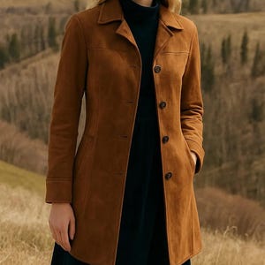 May include: A brown suede coat with a classic design. The coat features a notched collar, button closure, and pockets. The long coat is knee-length and has long sleeves. The coat is worn over a dark blue dress.