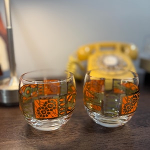 May include: Two clear, round drinking glasses with an orange and green geometric pattern. The glasses are on a dark wooden surface. A yellow rotary phone is in the background.