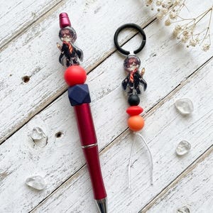 May include: A burgundy pen and a keychain with a Harry Potter theme. The pen has a red barrel and is adorned with beads and a small Harry Potter figure. The keychain has a black ring and a similar Harry Potter figure and bead design.