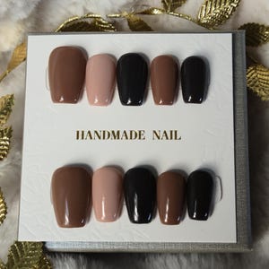 Peut inclure: Un ensemble de dix faux ongles de différentes couleurs neutres, dont des nuances de marron, de nude et de noir. Les ongles sont présentés sur une carte blanche avec le texte "HANDMADE NAIL". Les ongles ont une finition brillante.