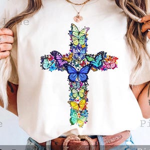 May include: Cream-colored t-shirt featuring a cross design made of colorful butterflies. The butterflies are in shades of blue, purple, yellow, and green. The text "Pixel Lane Market" and "Digital Download" are visible.