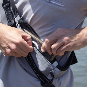 May include: A gray and black fanny pack with a black strap is being unzipped. The pack has a zippered pocket and a white ring pull. A black watch is on the person's wrist.