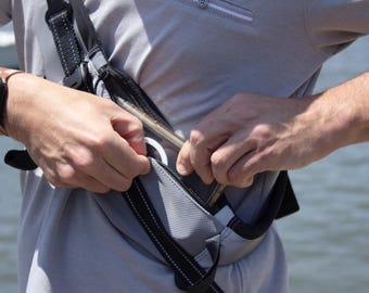 May include: A gray and black fanny pack with a black strap is being unzipped. The pack has a zippered pocket and a white ring pull. A black watch is on the person's wrist.