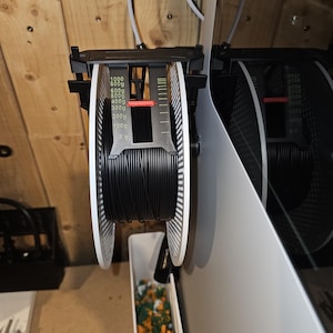 Könnte beinhalten: Eine schwarze Filamentrolle für den 3D-Druck, montiert auf einem weiß-schwarzen Halter. Der Halter hat eine Gewichtsanzeige in Gramm. Die Spule ist teilweise abgewickelt, mit einem weißen Behälter darunter, der mit farbigem Material gefüllt ist.