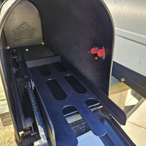 May include: A black metal letterbox with an open door, showing an interior tray and a red flag. The letterbox is mounted on a wooden post. The brand name is visible on the inside of the letterbox.