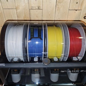 May include: A black 3D printer filament storage unit with four spools of filament in white, blue, yellow, and red. The unit has a clear, curved top and the text "Bambu Lab AMS 2" is visible on the front.