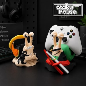 May include: Two snail-shaped controller stands. The left one is tan with black markings and a yellow shell. The right one is tan with a green base, holding a white game controller, and features three miniature swords across its chest.