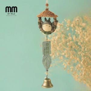 Puede incluir: Un carillón decorativo con una figura de Totoro en una casa con techo de paja. El carillón tiene un colgante en forma de hoja con texto japonés y una pequeña campana de latón. El diseño incluye el texto "MM BY MILA".