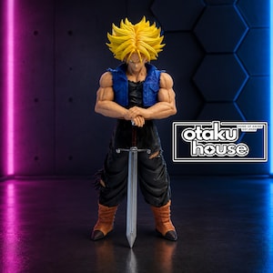 Op de afbeelding: Een verzamelbaar anime-figuur met stekelig geel haar, gekleed in een blauw vest over een zwart shirt en een zwarte broek met gescheurde knieën. Het figuur staat met de handen op het gevest van een lang zilveren zwaard. Tekst leest otaku house.