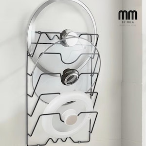 May include: Black metal pot lid organizer holding three glass and metal lids. The organizer is mounted on a white wall. The lids have silver and black handles. The organizer has the text "mm BY MILA DESIGNED BY MILA - EST 2020".
