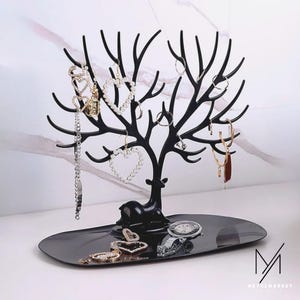 Puede incluir: Organizador de joyas de metal negro con forma de árbol con base de ciervo. El árbol sostiene collares, pulseras y pendientes. Una bandeja en la base contiene joyas adicionales y un reloj. El organizador está sobre una superficie blanca.