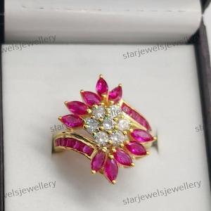 Könnte beinhalten: Ein Goldring mit einer Anordnung runder weißer Steine in der Mitte, umgeben von rosafarbenen Steinen im Marquise-Schliff in einem Blumenmuster, mit zusätzlichen rosafarbenen Steinen im Baguette-Schliff am Ringband, in einer dunklen Schmuckschatulle.