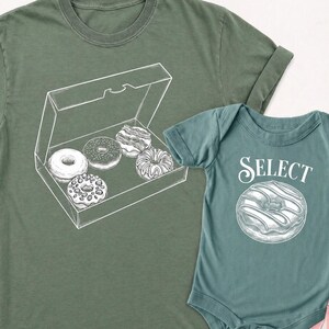 May include: Olive green t-shirt with a white illustration of a donut box containing five donuts. A matching teal baby onesie features a donut graphic and the word "SELECT".