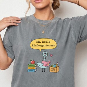 Puede incluir: Una camiseta gris carbón de manga corta con una paloma de dibujos animados leyendo un libro. Un bocadillo amarillo arriba dice: "Oh, hello kindergarteners!". Junto al ave hay pilas de libros etiquetados "Adventures", "Laughs" y "Dreams", además de una caja con "Good Ideas", un lápiz y una manzana roja.