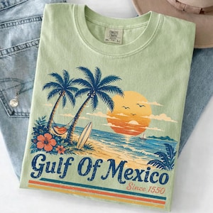 Puede incluir: Una camiseta verde claro con un gráfico de una escena de playa con palmeras, una hamaca, una tabla de surf y una puesta de sol. El texto dice Gulf Of Mexico Since 1550. La camiseta está sobre unos vaqueros azules junto a un sombrero beige de ala ancha.