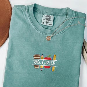 Può includere: Una maglietta Comfort Colors verde acqua con un design ricamato a tema insegnante. Il design include una tazza di caffè, una matita, una mela, un righello e il nome "Ms. Carter" in bianco. Una collana d'argento con un ciondolo è drappeggiata sulla maglietta.