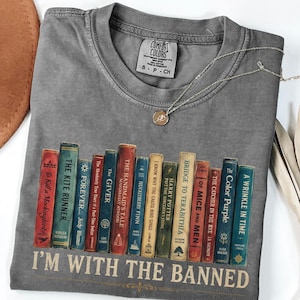Könnte beinhalten: Ein anthrazitfarbenes Kurzarm-T-Shirt mit einer Grafik verschiedener Buchrücken mit Titeln wie The Giver und The Handmaid's Tale sowie dem darunter gedruckten Text I'M WITH THE BANNED. Das Shirt hat einen Rundhalsausschnitt und ein Comfort Colors-Etikett.