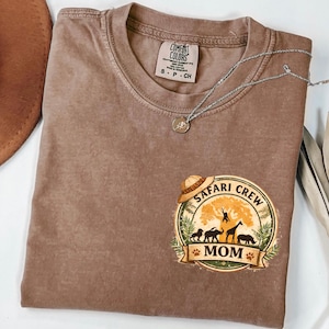 May include: A brown Comfort Colors t-shirt with a "Safari Crew Mom" graphic. The graphic features a safari hat, animals, and the word "Mom". A silver necklace with a pendant is draped across the shirt.