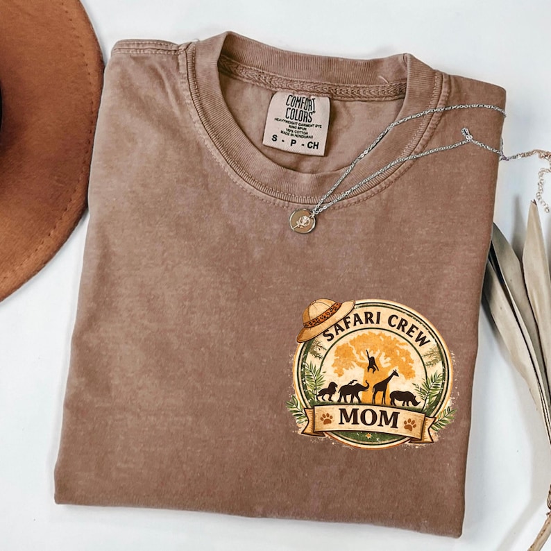 May include: A brown Comfort Colors t-shirt with a "Safari Crew Mom" graphic. The graphic features a safari hat, animals, and the word "Mom". A silver necklace with a pendant is draped across the shirt.