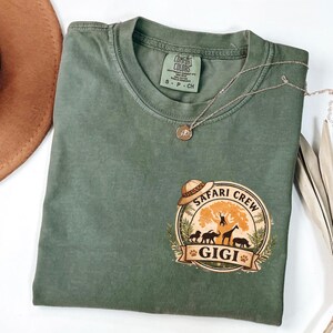 May include: Olive green t-shirt with a round graphic that says "Safari Crew" and "GIGI". The graphic features a safari hat, animals, and a tree. A silver necklace with a pendant is draped across the shirt.