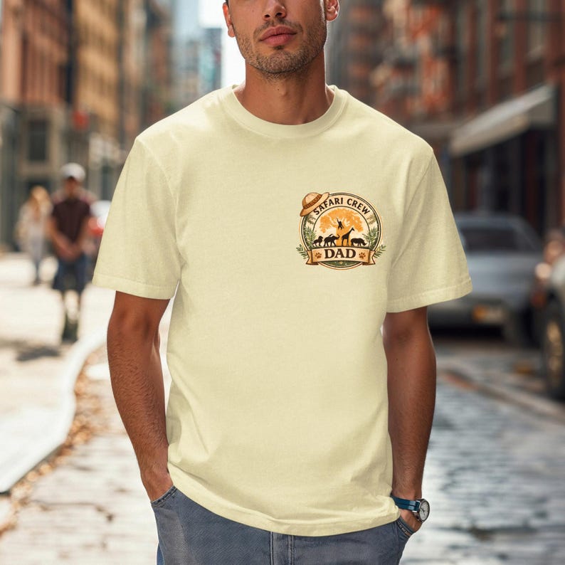 May include: Cream-colored t-shirt with a circular graphic featuring a safari theme. The graphic includes a hat, animals, and the words "Safari Crew Dad". The shirt has short sleeves and a crew neck.