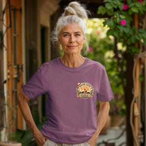 May include: A mauve t-shirt featuring a circular graphic with the words "Safari Crew" and the name "Gigi". The graphic includes silhouettes of animals and a safari hat. The shirt is short-sleeved.