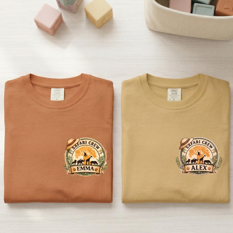 May include: Two short-sleeved t-shirts, one in burnt orange and the other in tan, each featuring a "Safari Crew" graphic with animal silhouettes and a name. The shirts are laid flat on a light wood surface.