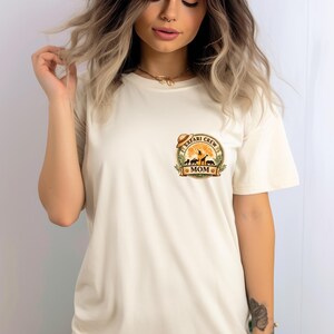 May include: Cream-colored t-shirt featuring a circular graphic with the words "Safari Crew Mom." The graphic includes silhouettes of animals like giraffes and zebras, a sun, and a safari hat. The shirt has short sleeves.