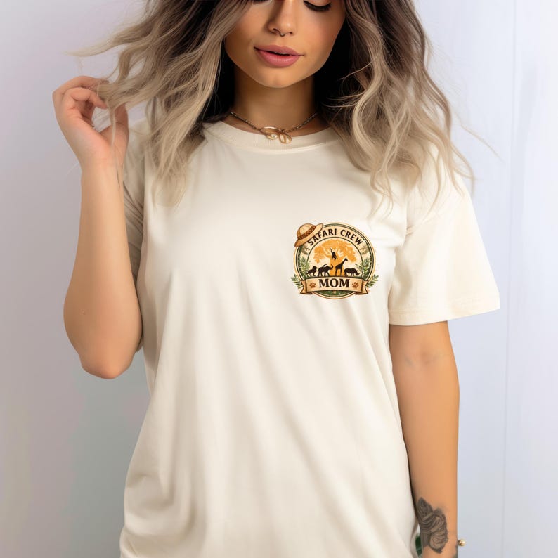 May include: Cream-colored t-shirt featuring a circular graphic with the words "Safari Crew Mom." The graphic includes silhouettes of animals like giraffes and zebras, a sun, and a safari hat. The shirt has short sleeves.