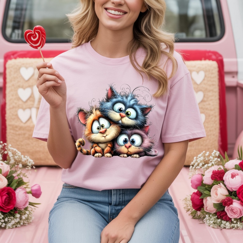 May include: A light pink t-shirt featuring a cartoon graphic of three adorable cats in orange, blue, and purple. The cats have large eyes and are smiling. The person is holding a heart-shaped lollipop.
