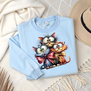May include: A light blue sweatshirt featuring a whimsical illustration of three cartoon cats with large eyes and bows. The cats are in shades of gray, blue, and orange. The sweatshirt is folded on a white surface.