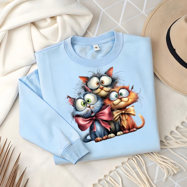May include: A light blue sweatshirt featuring a whimsical illustration of three cartoon cats with large eyes and bows. The cats are in shades of gray, blue, and orange. The sweatshirt is folded on a white surface.