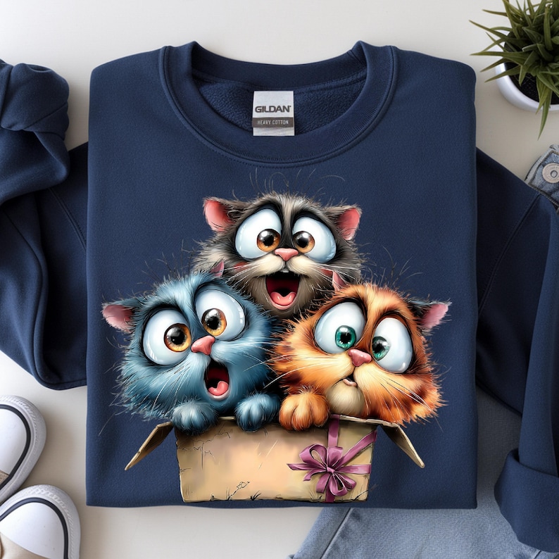 May include: Navy blue sweatshirt featuring a cartoon illustration of three surprised cats peeking out of a cardboard box. The cats have large eyes and expressive faces, with one cat's mouth open in a surprised expression. The box is tied with a pink ribbon.