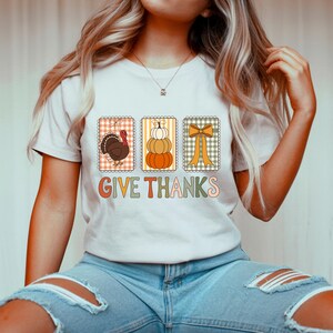 May include: A pale t-shirt with a Thanksgiving graphic. The design features a turkey, pumpkins, and a bow, each in decorative frames. The words "GIVE THANKS" are printed in a retro font with orange, green, and pink colours.
