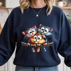 May include: Navy blue sweatshirt featuring a whimsical illustration of three cartoon cats perched on a branch. The cats have large, expressive eyes and are in shades of orange, purple, and blue. The sweatshirt is made of a soft material.