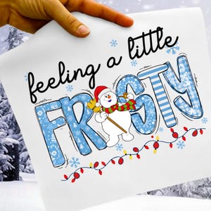 Op de afbeelding: Een witte print met de tekst "feeling a little frosty" in zwart script. Het woord "frosty" is blauw met sneeuwvlok- en streepjespatronen. Een sneeuwman met een hoed en sjaal houdt een bezem vast. Een slinger van lichtjes staat onderaan.