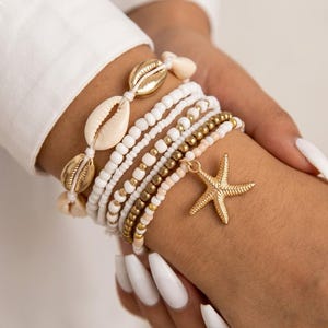 Könnte beinhalten: Eine Sammlung von Armbändern mit Perlen in Weiß, Gold und Pfirsichtönen. Ein Armband hat einen goldenen Seestern-Anhänger. Weitere Armbänder enthalten goldene Perlen und natürliche Muscheln.