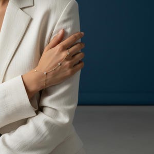 Puede incluir: Una delicada cadena de mano dorada con pequeñas y brillantes piedras preciosas. La cadena conecta un anillo con una pulsera, con una serie de piedras preciosas y pequeñas cuentas doradas. La joya se muestra sobre un fondo azul y una blazer blanca.