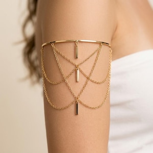 May include: Gold-toned arm cuff featuring a delicate design. The upper band encircles the arm, with multiple chains cascading down. Vertical bar pendants are evenly spaced along the chains. This arm cuff is a stylish accessory.