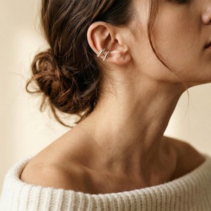 May include: Silver ear cuffs, featuring a minimalist design, are worn on the ear. The jewelry is visible against the skin. The person is wearing a white sweater. The hair is pulled back in a bun.