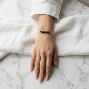 May include: A gold-toned bracelet featuring a string of dark, irregularly shaped beads. The bracelet is worn on a wrist, set against a backdrop of white fabric and a marble surface. The beads appear to be a dark blue or black color.