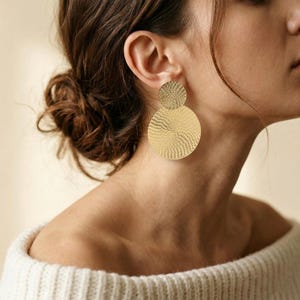 Könnte beinhalten: Goldfarbene, strukturierte Scheibenohrringe. Die Ohrringe bestehen aus einer kleineren, kreisförmigen Scheibe über einer größeren, kreisförmigen Scheibe. Das Design hat ein Spiralmuster. Die Person trägt einen weißen Pullover.