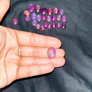 Könnte beinhalten: Eine Sammlung ovaler, polierter Edelsteine in Lila- und Rosatönen. Ein Edelstein liegt in einer offenen Hand und zeigt seine Farbvariationen. Die Edelsteine sind auf einer dunklen Oberfläche angeordnet.