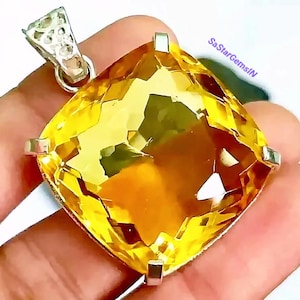 Puede incluir: Un gran colgante de citrino amarillo con corte cuadrado y facetas, montado en una base de metal plateado con garras y una anilla triangular texturizada. El texto SaStarGemsIN está impreso en violeta en la parte superior derecha.