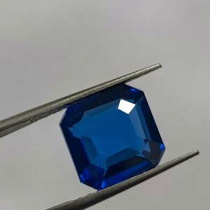 Könnte beinhalten: Ein facettierter, rechteckiger, tiefblauer Edelstein wird von einer silbernen Pinzette gehalten. Der Stein hat einen Brillantschliff und reflektiert das Licht. Der Hintergrund ist ein weiches, neutrales Grau.