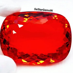 Könnte beinhalten: Ein großer, leuchtend roter Edelstein mit Kissenschliff und facettierter Oberfläche, die das Licht reflektiert. Der Stein weist eine brillante, gesättigte karmesinrote Farbe auf. Der Text SaStarGemsIN ist in Schwarz über dem Edelstein aufgedruckt.