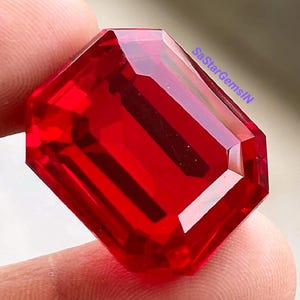 以下が含まれることがあります： 鮮やかな透明感のある赤色のエメラルドカットの宝石で、光を反射する複数のファセットが特徴です。石は深い深紅の色合いをしており、右上に小さな青い文字でSaStarGemsINというテキストが記されています。