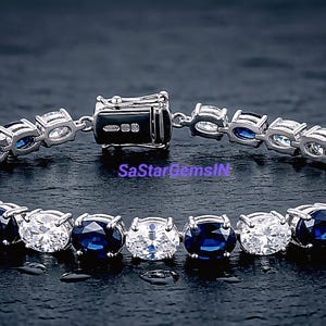 Könnte beinhalten: Ein silbernes Armband mit abwechselnd weißen und tiefblauen Edelsteinen. Das Armband wird mit einem Verschluss gesichert und auf einer dunklen, strukturierten Oberfläche präsentiert. Der Text SaStarGemsIN ist sichtbar.
