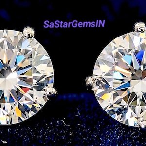 Könnte beinhalten: Ein Paar funkelnde Diamant-Ohrstecker. Jeder Ohrring hat einen großen, runden Diamanten im Brillantschliff, der in einer silberfarbenen Krappenfassung gefasst ist. Die Diamanten reflektieren das Licht und erzeugen ein schillerndes Funkeln.